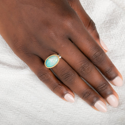 An oval ring featuring a light blue-green opal stone in a gold setting, with a silver-toned band, shown on a person's finger.