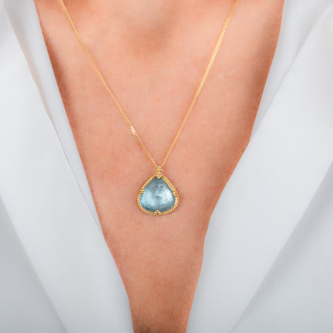 A gold necklace with a teardrop-shaped aquamarine gemstone pendant, shown worn on a model's neck.