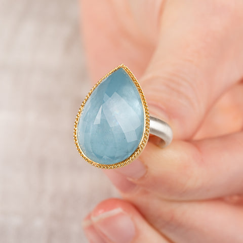 Close up of a hand holding an aquamarine ring with gold bezel and sterling silver band on white background