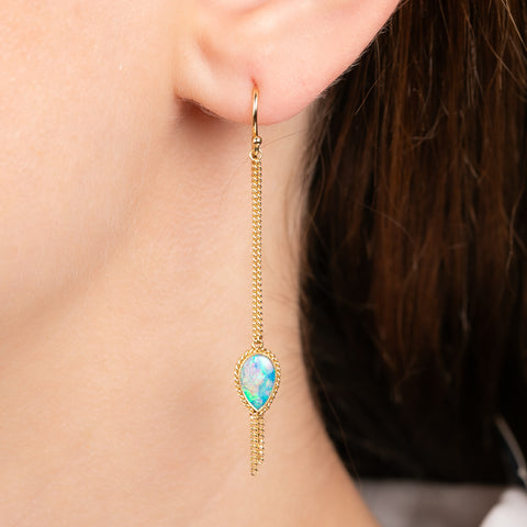 A gold drop earring with a teardrop-shaped black opal stones, hanging from a delicate chains, shown worn on a model's ear.