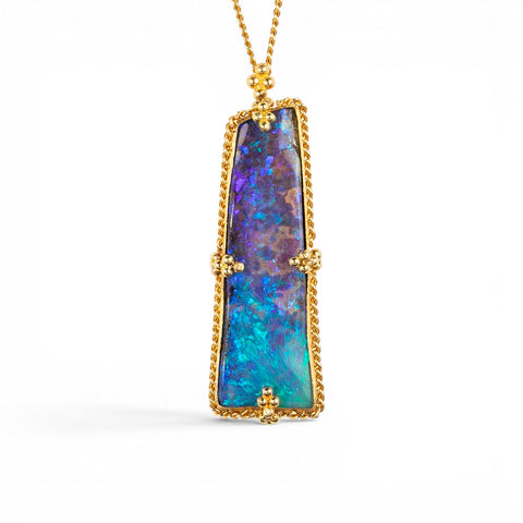 A gold pendant necklace featuring a vertically oriented, rectangular iridescent boulder opal stone with blue and purple play-of-color, set in a textured gold bezel, suspended from a gold chain against a white background.