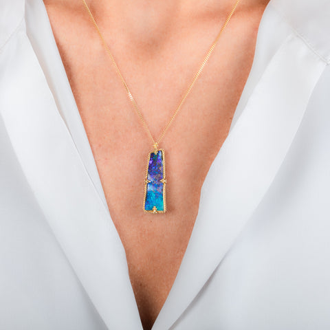 A gold pendant necklace featuring a boulder opal stone with blue and purple set in a textured gold bezel, worn on a person's neck.