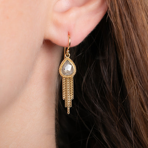 A close-up of a gold dangle earring with a faceted teardrop-shaped diamond and delicate gold chain fringe, worn on a person's ear.