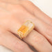 A gold ring featuring a large, rectangular, Citrine gemstone set in a detailed gold bezel, worn on a person's finger.