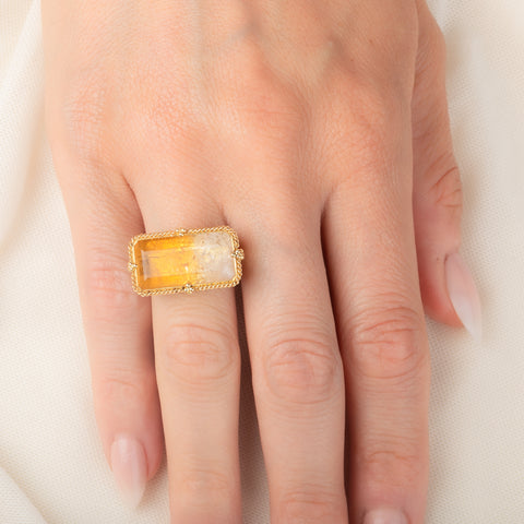 A gold ring featuring a large, rectangular, Citrine gemstone set in a detailed gold bezel, worn on a person's finger.