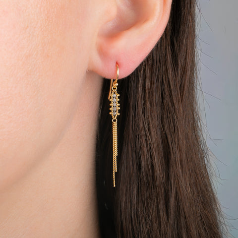 Gold earrings with gold tassels dangling between rows of four silver diamonds, shown in a person's ear.
