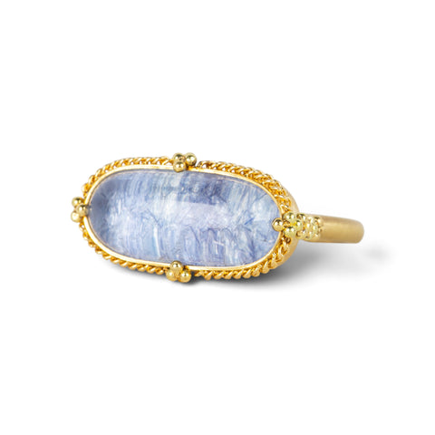An oval ring featuring a textured light blue dumortierite stone in a yellow gold bezel setting and a matching gold band, shown on a white background.