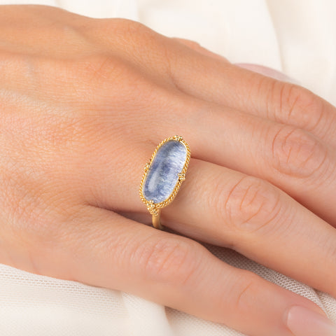 An oval ring featuring a textured light blue dumortierite stone in a yellow gold bezel setting and a matching gold band, worn on a person's finger.