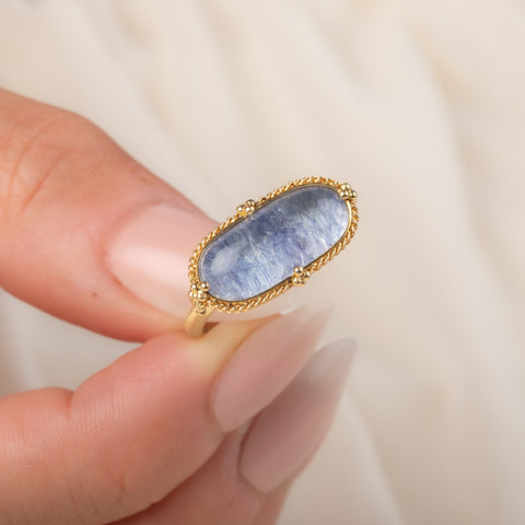 An oval ring featuring a textured light blue dumortierite stone in a yellow gold bezel setting and a matching gold band, held in hand.