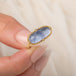 An oval ring featuring a textured light blue dumortierite stone in a yellow gold bezel setting and a matching gold band, held in hand.