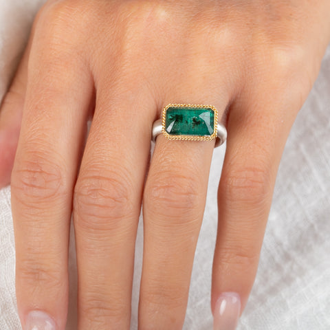 A rectangular emerald stone ring with a gold bezel and a sterling silver band, worn on a finger