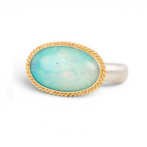 An oval ring featuring a light blue-green opal stone in a gold setting, with a silver-toned band, shown on a plain white background.