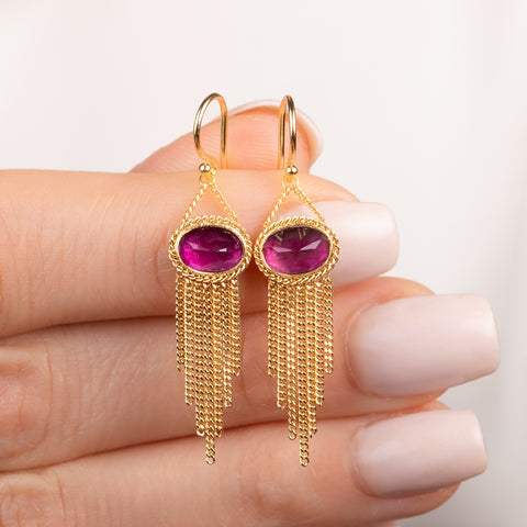 A pair of gold dangle earrings with oval pink fluorite gemstones and cascading chain tassels, held in hand.