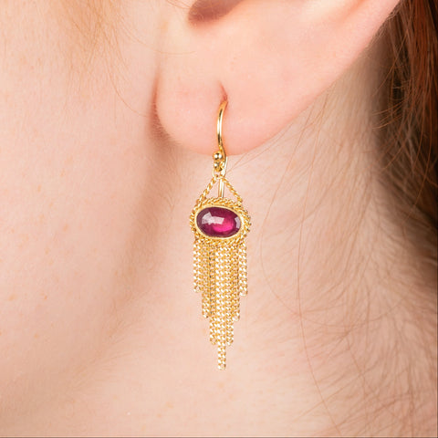 Gold earrings featuring oval rhodolite garnet gemstones, shown worn in a person's ear.