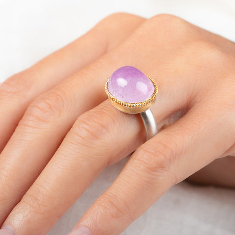 A close-up of a hand wearing a gold and silver ring with a smooth oval kunzite stone.