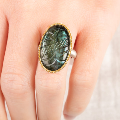 A close up of a person wearing a handmade fine jewelry labradorite ring with carved design set in 18k gold with a sterling silver band