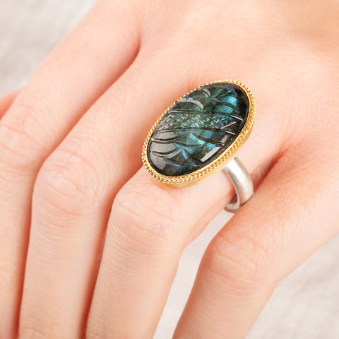 A close up of a person wearing a handmade fine jewelry labradorite ring with carved design set in 18k gold with a sterling silver band