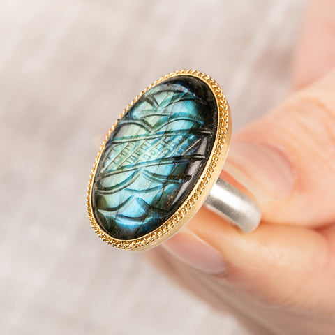 A close up of a person holding a handmade fine jewelry labradorite ring with carved design set in 18k gold with a sterling silver band