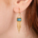 Gold earrings featuring London blue topaz gemstones and gold tassels, shown worn on a person's ear.