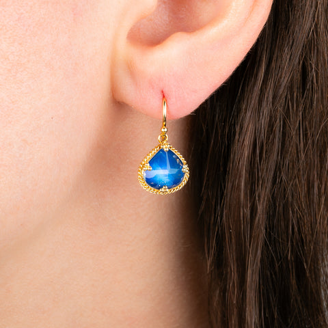 A close-up of a person's ear wearing a gold teardrop earring with a bright blue moonstone.