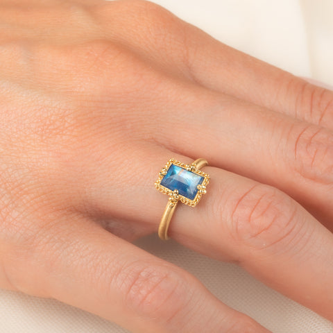 A gold ring featuring a rectangular moonstone set in a 18k gold bezel, worn on a person's finger.