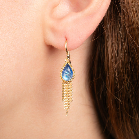 A close-up of a person's ear wearing a gold dangle earring with a teardrop-shaped blue moonstone and a gold chain tassel.