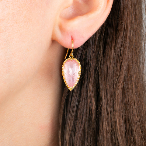 A gold drop earring featuring a pink morganite gemstone in a textured gold bezel setting, shown worn in a person's ear.