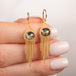 A pair of gold dangle earrings with oval diamond gemstones and cascading gold chain fringe, show held in hand.