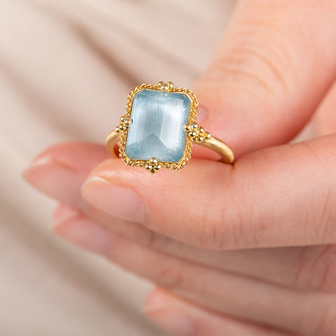 Gold ring with a blue aquamarine gemstone held in a hand against a beige background