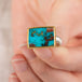 Close up of a hand holding a turquoise ring with gold bezel and silver band