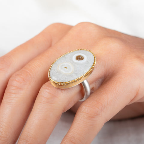 An oval ring featuring a solar quartz stone set in a textured gold bezel with a sterling silver band, worn on a person's finger.