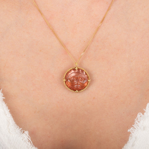 Gold necklace with a circular sunstone pendant featuring a sun face design on a person's neck