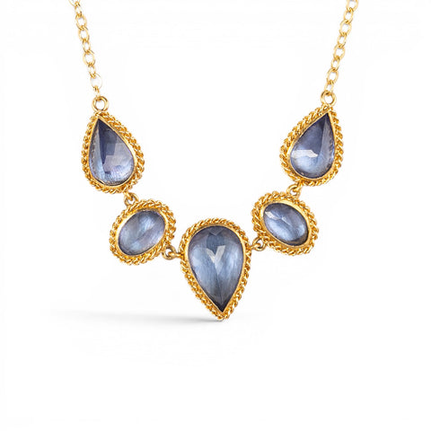 A gold necklace featuring five tanzanite gemstones in textured gold bezels, displayed on a clean white background.