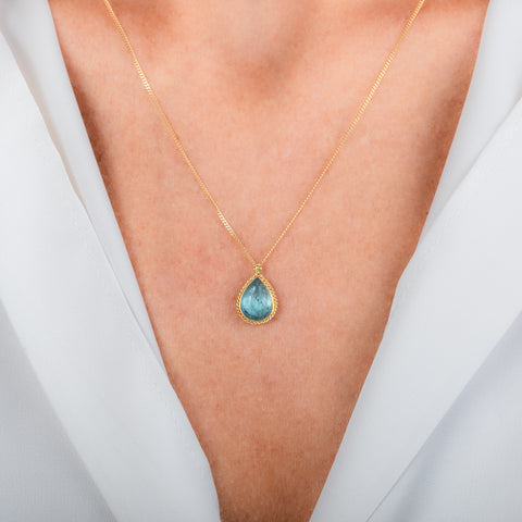 A gold necklace featuring a teardrop-shaped aquamarine necklace, shown worn on a model's neck.