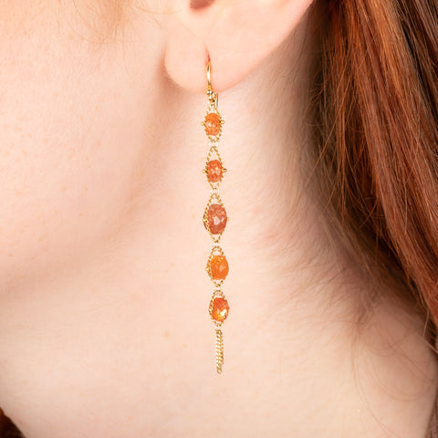 Gold earrings featuring rows of orange sunstones, worn in a person's ear.