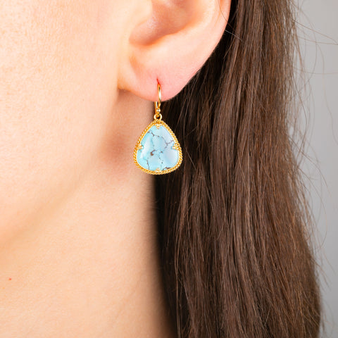 A close-up of a person's ear wearing a teardrop-shaped drop earring with a light blue turquoise stone and a gold bezel setting.