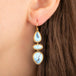 A close-up of a person's ear wearing a gold drop earring with three stacked turquoise-blue stone settings.
