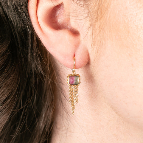 A person wears gold earrings with watermelon tourmaline gemstones and gold chain tassels