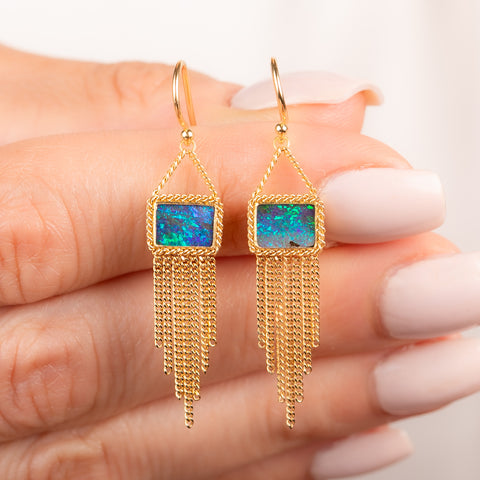 A pair of gold drop earrings featuring rectangular boulder opal stones and cascading gold fringe, shown held in hand.