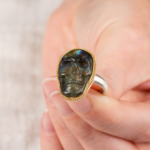 A hand holds a labradorite skull ring set in an 18k gold bezel with a sterling silver band