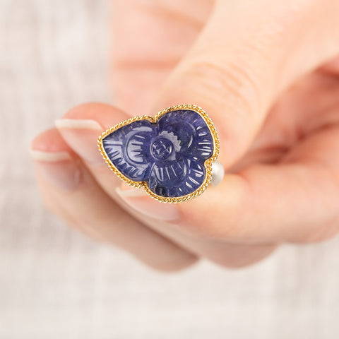 A close up of a person holding a handmade gemstone ring with carved tanzanite stone set in gold and complete with sterling silver band