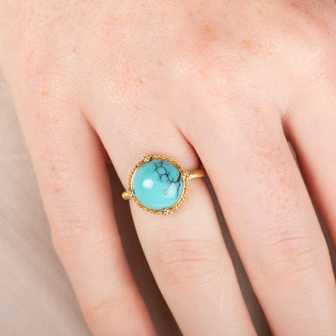 Gold ring with a turquoise stone on a person's finger against a beige background