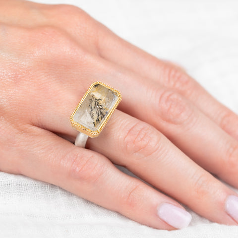 Gold and silver ring with a rectangular, dendritic quartz stone on a hand against a white background