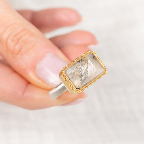 Gold and silver ring with a dendritic quartz stone held by a hand on a light background