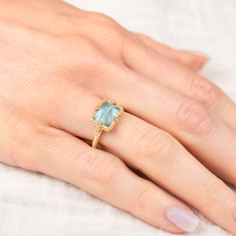 Gold ring with a light blue aquamarine gemstone on a hand against a white background