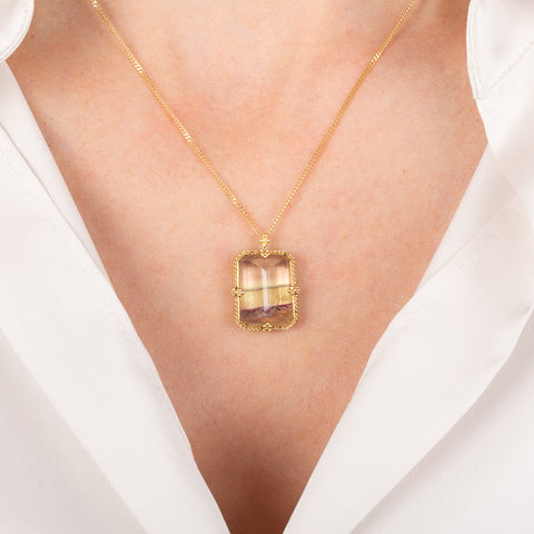 Gold necklace with a rectangular fluorite gemstone worn by a person in a white outfit.