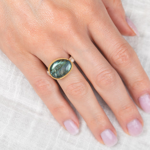 Hand wearing a gold ring with a labradorite stone on a white fabric background