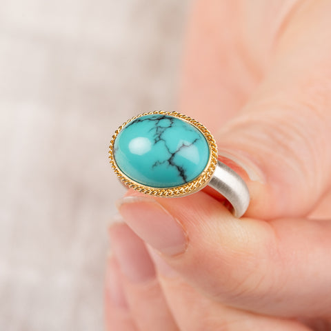 Close up of a hand holding a turquoise ring with gold bezel and sterling silver band on white background