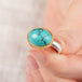 Close up of a hand holding a turquoise ring with gold bezel and sterling silver band on white background