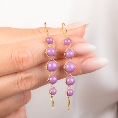 A close-up of a hand holding a pair of gold drop earrings with purple phosphosiderite stones, against a soft white background.